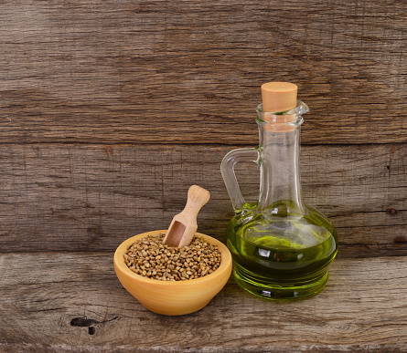 Eine gläserne Flasche mitgrünen Hanföl steht neben einer Holzschale, die mit Hanfsamengefüllt ist und in der ein Löffel steckt.
